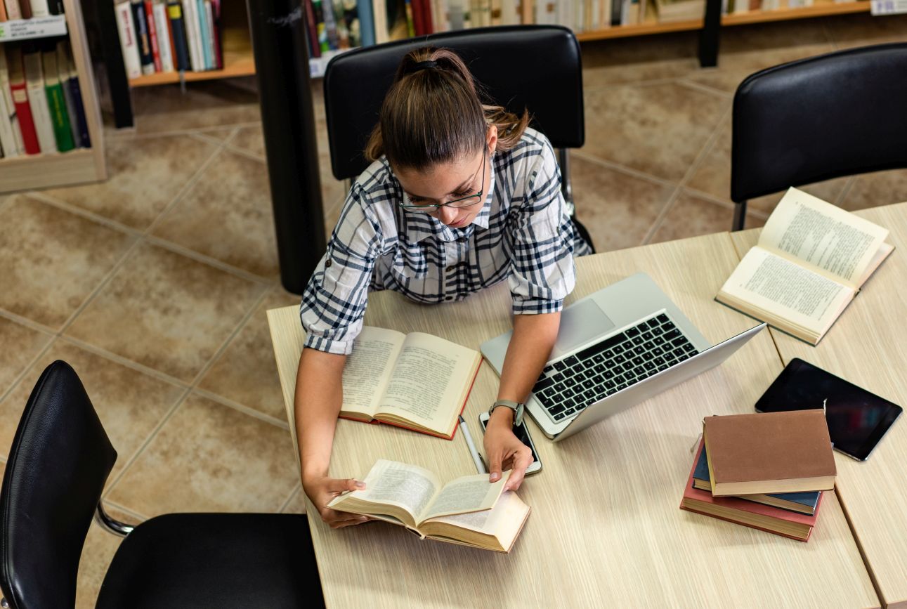 Eine Person sitzt in einer Bibliothek am Tisch, liest in mehreren geöffneten Fachbüchern und arbeitet parallel am Laptop. Szene zeigt sorgfältige Informationsrecherche.